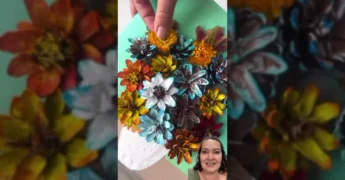 Imagem de uma obra de arte 3D feita com pinhas pintadas em diversas cores, formando um vaso de flores em um painel, com detalhes de folhas e flores desidratadas - Kidica10 | Mel Kiyota