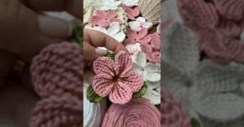 Uma mulher crocheta uma flor de barbante rosa, com muitas flores de crochê rosa e branco espalhadas sobre uma superfície. Créditos: Geise Crochê Criativo / Instagram